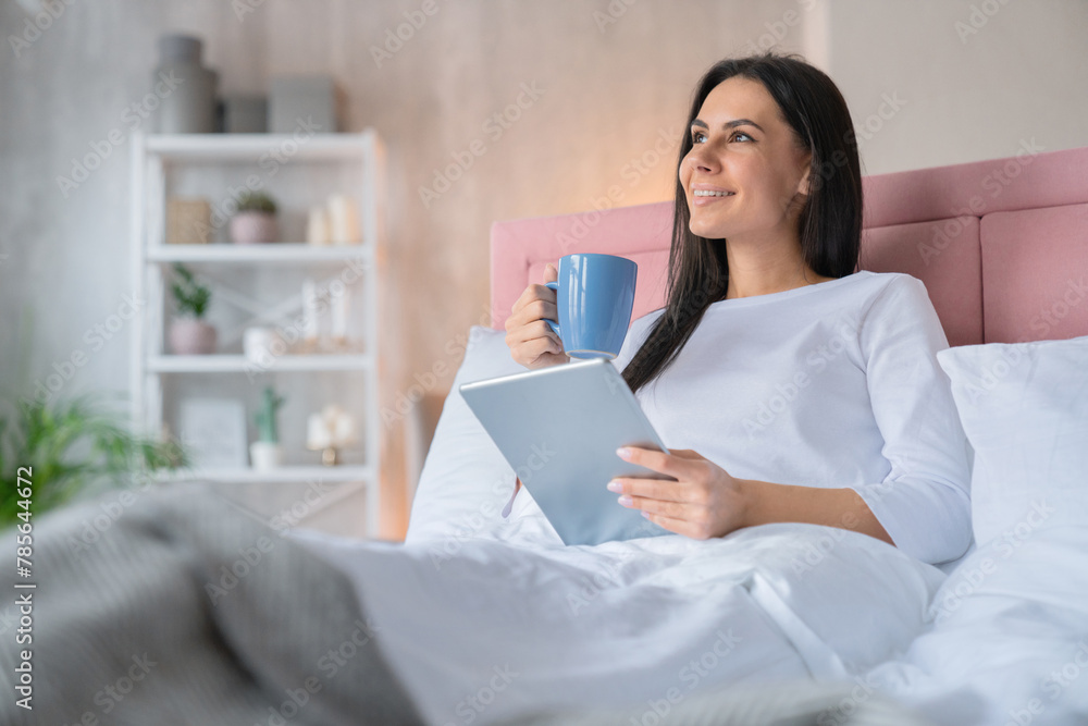 Dreaming moment. Side view shot of young beautiful woman with brown hair in light white pajamas in home bed with cup of morning coffee tea and tablet pad in hands Dreaming moment. Side view shot of young beautiful woman with brown hair in light white pajamas in home bed with cup of morning coffee tea and tablet pad in hands