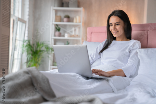 Attractive smiling caucasian woman working remotely on notebook laptop while being lying at home bed. Woman sitting with computer  studying at home  e-learning and remote freelance job concept
