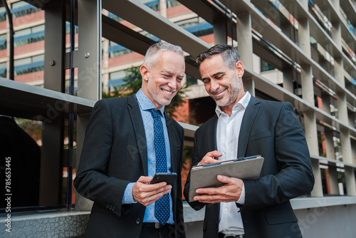 Two professional colleagues suited up and engrossed, collaborate seamlessly as they navigate data and charts on a sleek tablet, their expressions a blend of focused determination and strategic insight