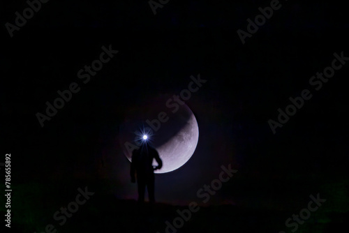 A stunning silhouette of a lone individual wearing a headlamp, standing before a backdrop of the crescent moon in a dark, serene night sky