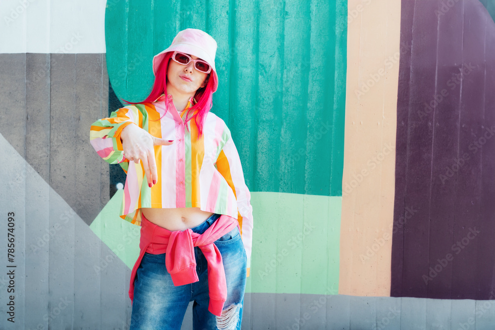 Young woman with pink hair and sunglasses in Bucket hat and multicolor strippled shirt posing on graffiti wall background. Street artist with mural, hipster, blogger. Urban street fashion - obrazy, fototapety, plakaty 