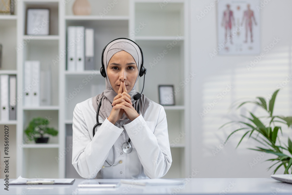 Focused female doctor wearing a hijab engaged in a serious video call, looking directly at the camera in a modern office setting. Focused female doctor wearing a hijab engaged in a serious video call, looking directly at the camera in a modern office setting.