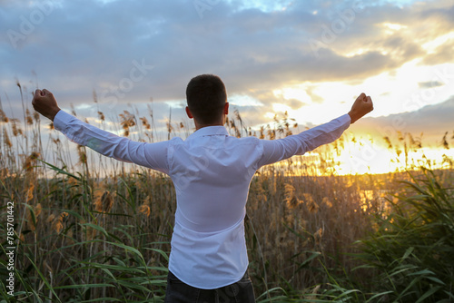 A man is standing in a field with his arms raised in the air, looking up at the sky. The sky is filled with clouds, and the sun is setting in the distance. Scene is one of joy and celebration
