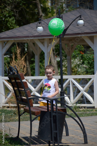 a boy with a green balloon stands near a bench in a city park. active lifestyle and walks in the fresh air. fun and entertainment for children