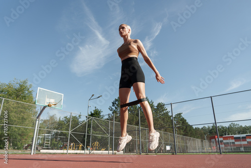 Focused athletic woman using resistance bands for leg workout on an outdoor basketball court with a clear sky background. © arthurhidden