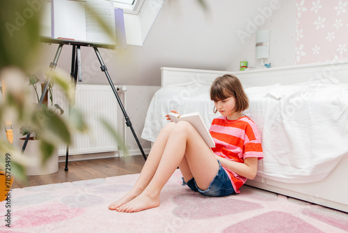 A teenage girl sits on the floor of her room and reads a book. Schoolchildren study at home. Reading books as a hobby