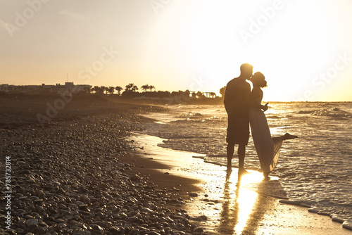 Silhouette young man, and young woman, loving young couple embraced enjoying a romantic walk at the beach on a hazy day at dusk, walking barefoot, getting wet, teasing and kissing one another.