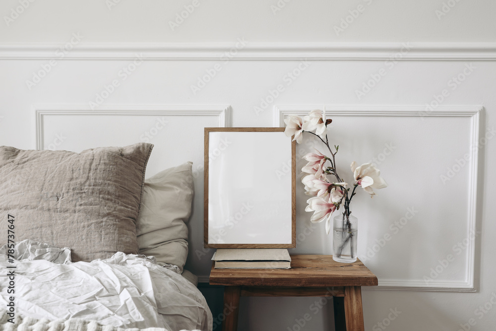 Blank wooden picture frame mockup on old book. Wooden night stand with fluted glass vase. Blooming magnolia tree branches. Scandinavian interior. Elegant bedroom. White wall background, stucco decor. - obrazy, fototapety, plakaty 