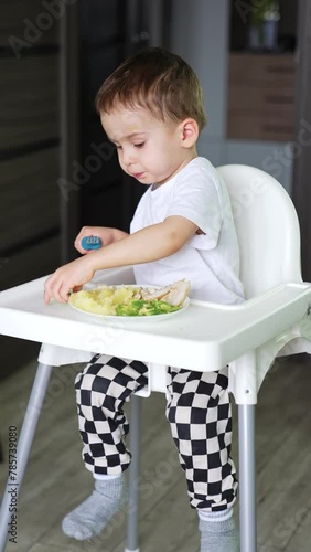 Little toddler boy sitting at high chair having meals. Kid holding a fork takes a piece of cucumber from the table and etas it. Vertical video. photo