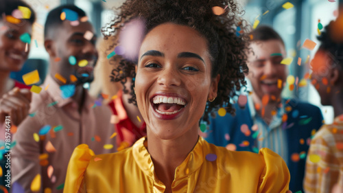 Radiant Woman Celebrating with Confetti and Friends. Joyful woman in yellow blouse with a wide smile, enjoying a confetti party with diverse colleagues in the office.
