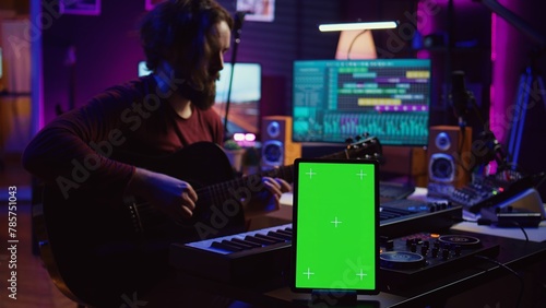 Composer performing acoustical guitar in his home studio, playing and practicing on strings. Musician learning multiple songs on instrument, uses tablet to run greenscreen display. Camera B.