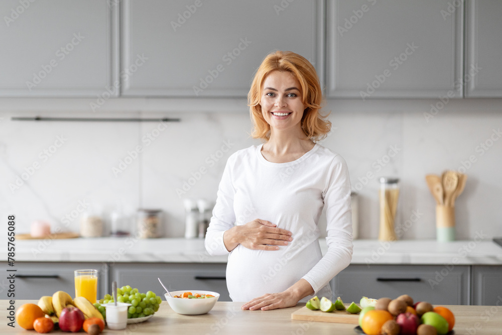 Cheerful pregnant woman standing in kitchen, cooking