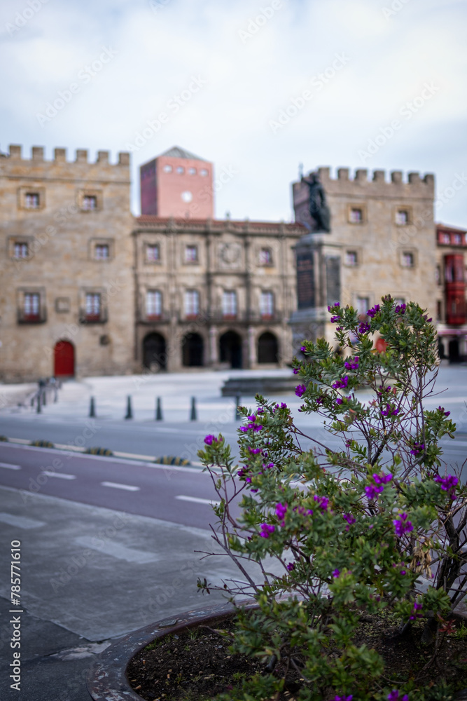 Purple flowers bloom, lush green leaves, historic Gijón plaza with castle, ancient architecture.