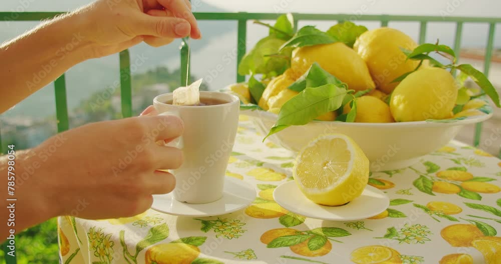 Tea bag steeps in a white mug on a table with lemons, mountains beyond. The act of dipping reflects a serene start to the day. Close-up of female hands. Tea bag steeps in a white mug on a table with lemons, mountains beyond. The act of dipping reflects a serene start to the day. Close-up of female hands.