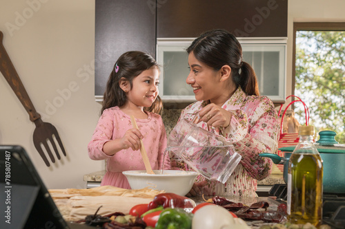 A Latina mother and daughter look happily into each other's eyes as the daughter mixes corn dough in a bowl to make tamales.