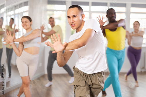 Expressive young Asian male instructor in white t-shirt and cargo pants leading modern dance exercise class for multiethnic group in well-lit fitness studio..