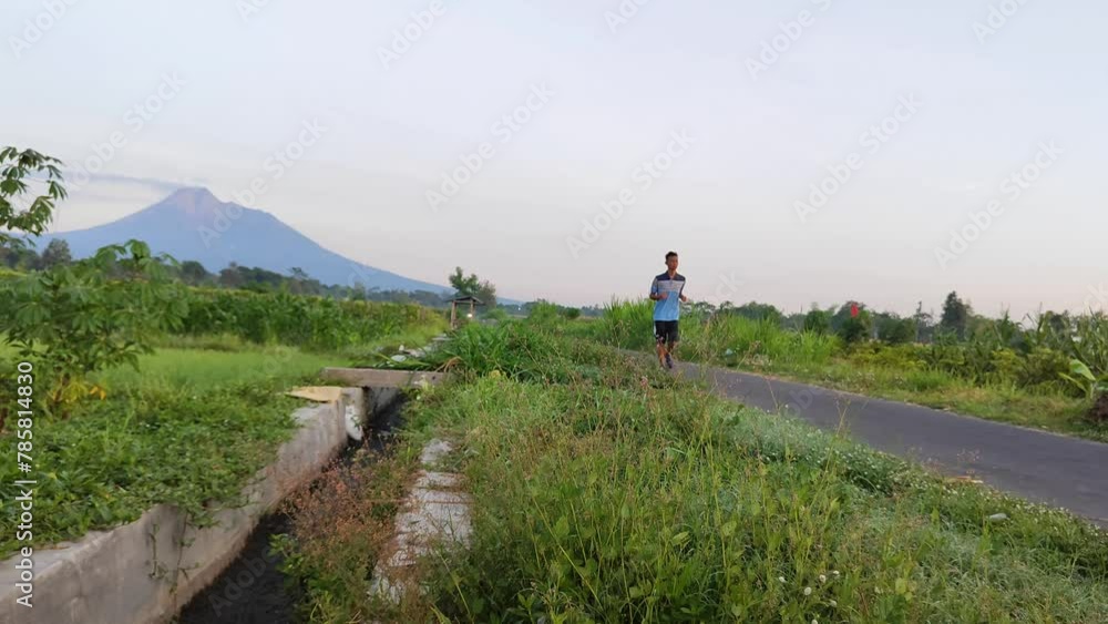 A man wearing a blue shirt and short pants is jogging on the road of the rice field area with the background of Mount Merapi A man wearing a blue shirt and short pants is jogging on the road of the rice field area with the background of Mount Merapi