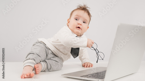 A cute child wearing a suit holds glasses and lies behind a laptop.
