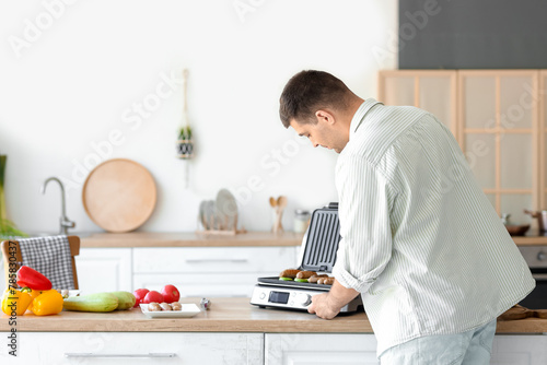 Young man cooking tasty sausages and vegetables on modern electric grill at table in kitchen