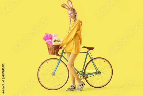 Young girl with bunny ears, Easter basket and bicycle on yellow background