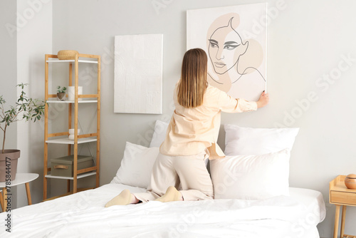 Young woman hanging stylish painting on light wall in bedroom, back view