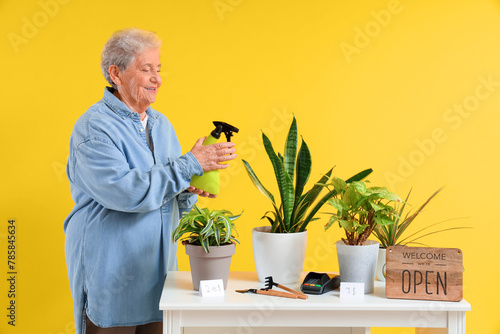 Senior florist with spray water bottle and plants on counter against yellow background