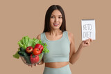 Beautiful young happy Asian woman holding notebook with text KETO DIET and bowl of fresh vegetables on brown background