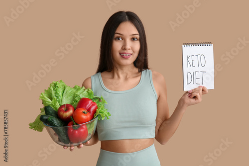Beautiful young happy Asian woman holding notebook with text KETO DIET and bowl of fresh vegetables on brown background © Pixel-Shot