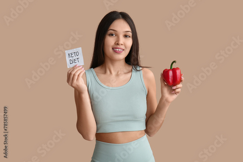Beautiful young happy Asian woman holding sticky note with word KETO and bell pepper on brown background © Pixel-Shot
