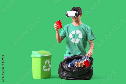 Tablou pe pânză Male ecologist in VR glasses with garbage bag and bin on green background