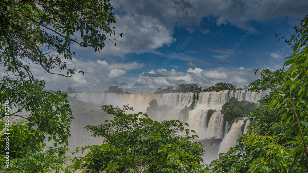 Beautiful waterfall landscape. Streams cascade down from the ledges of the rocks. Splashes, fog. Clouds in the blue sky. Lush tropical vegetation. Iguazu Falls. Argentina.