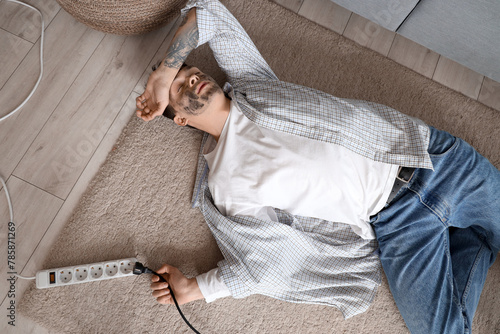 Obraz na plátně Electrocuted young man with burnt face and plug lying on floor at home, top view