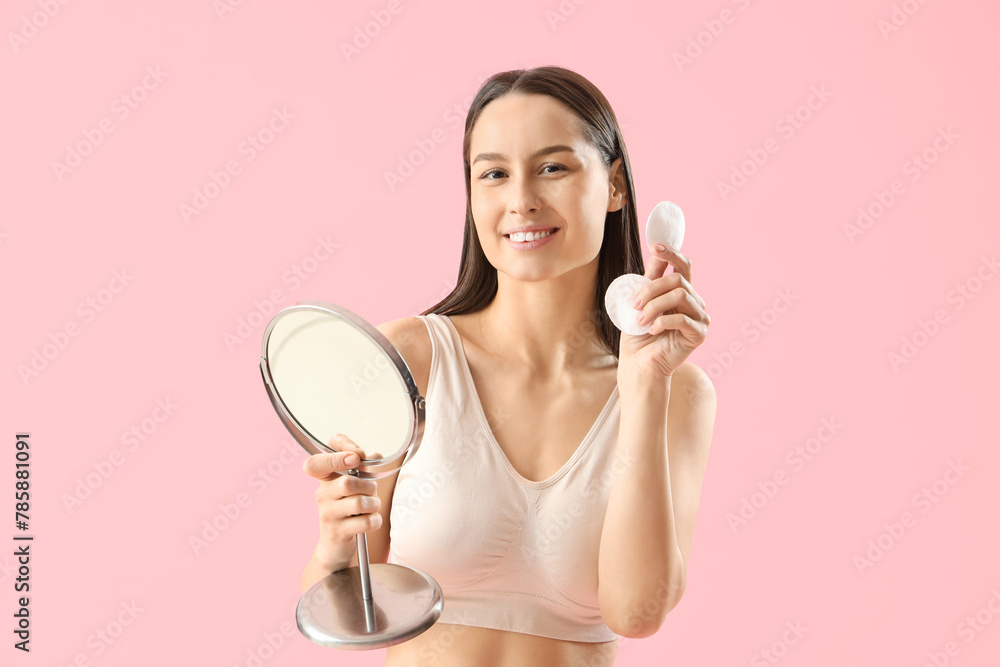 Young woman with cotton pads and mirror removing makeup on pink background Young woman with cotton pads and mirror removing makeup on pink background