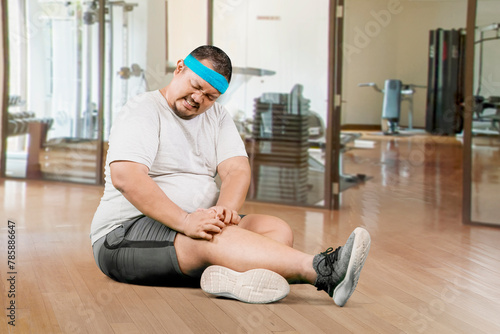Portrait of young plus size overweight man holding a knee suffering from an injury in a gym