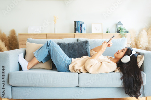 Happy young asian woman relaxing at home. Female smile sitting on sofa and holding mobile smartphone. Girl using video call to friend. Listening to music © Monster Ztudio