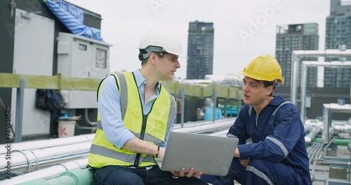 engineers manager and worker sitting on rooftop review plans on a laptop while on a construction site with high-rise buildings, indicating their different roles and responsibilities on the project