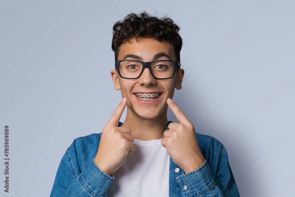 Dental dent care ad concept image - black сurly haired funny young man wear metal braces, eye glasses, show point white teeth smile. Isolated grey gray studio wall background. Positive optimistic. - obrazy, fototapety, plakaty 