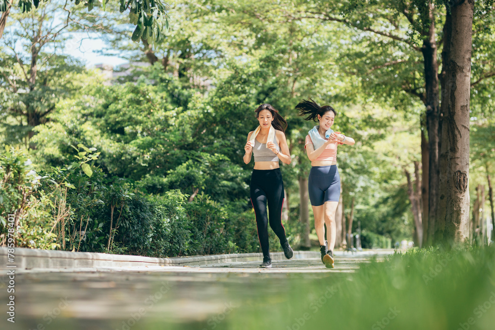 two Asian women engaging in physical activity within an urban park. They are incorporating smart devices to monitor and track their body data using technology.