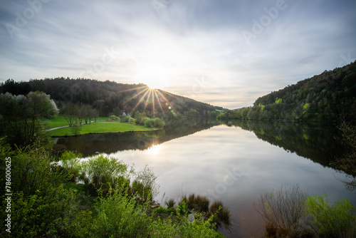 A lake in a landscape shot. A sunset and the natural surroundings are reflected in the water of the reservoir. Marbachstausee, Odenwald, Hesse