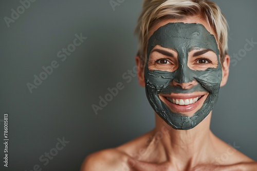 Portrait of a middle-aged woman with short blonde hair wearing a gray clay mask smiling at the camera on a gray background, studio shot photo