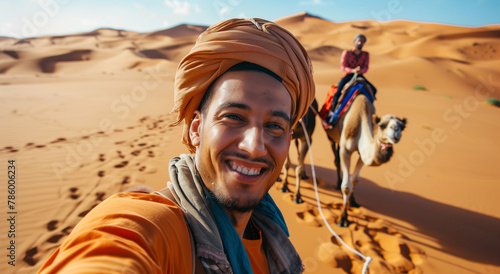 A man taking a selfie in the desert with a group of people on camels behind him  a happy and smiling man with a headscarf
