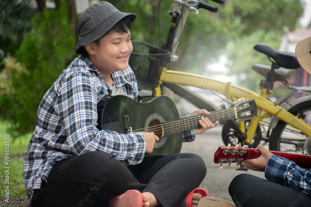 Obraz premium Asian boys hold guitar and playing near their bikes which parked by the narrow street in school area during their summer holiday, soft focus, summer activity of children concept.