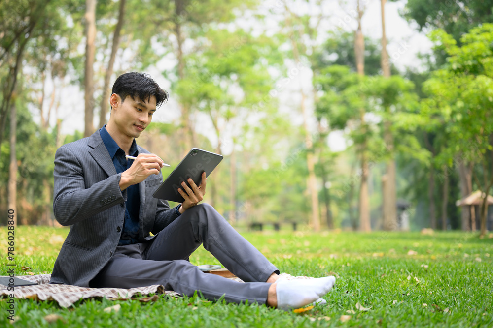 Full length of young businessman using digital tablet sitting on grass at park Full length of young businessman using digital tablet sitting on grass at park