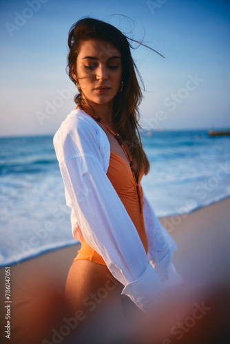 Pensive woman dressed in a white shirt and beachwear by the ocean at dusk