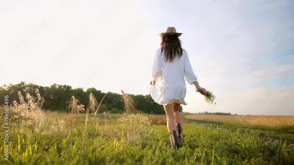 Brunette woman in airy white dress, sun hat and rubber boots with bouquet of field flowers walking along country road between agricultural fields in village, back view. Carefree, village life. Brunette woman in airy white dress, sun hat and rubber boots with bouquet of field flowers walking along country road between agricultural fields in village, back view. Carefree, village life.