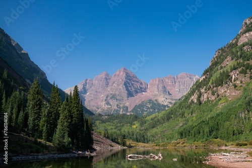 Image of a river and mountains with red tops behind the green range of trees under the blue sky. © Wirestock