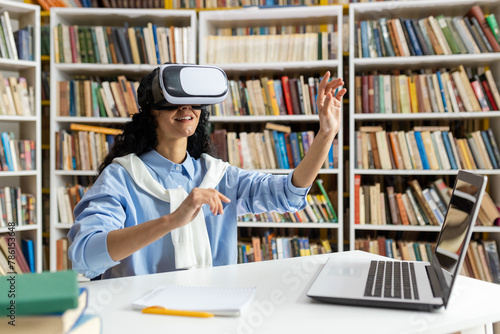 A young woman engages with a virtual reality headset, exploring digital worlds while surrounded by books in a library setting. This image captures her fascination and curiosity.
