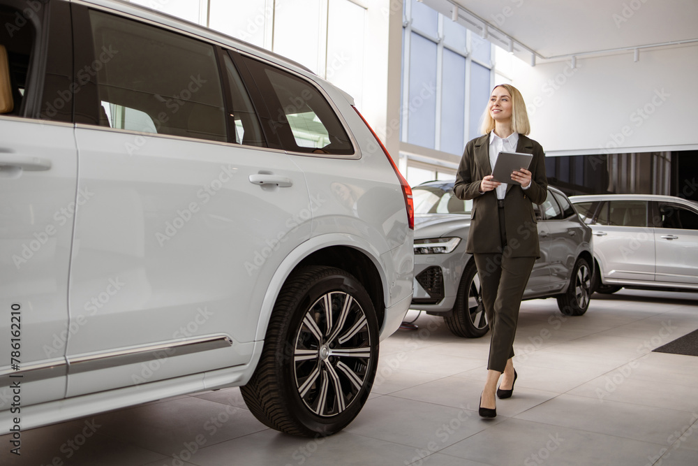 Professional Caucasian female dealer with digital tablet smiling and looking at camera friendly. Young saleswoman standing near modern automobile in bright car showroom. Professional Caucasian female dealer with digital tablet smiling and looking at camera friendly. Young saleswoman standing near modern automobile in bright car showroom.