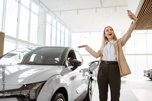 Successful businesswoman taking selfie using smartphone in car service. Happy smiling Caucasian girl buyer talking online about buying a new car.