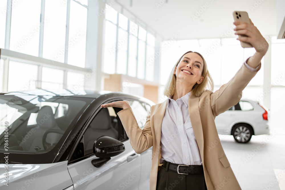 Successful businesswoman taking selfie using smartphone in car service. Happy smiling Caucasian girl buyer talking online about buying a new car. Successful businesswoman taking selfie using smartphone in car service. Happy smiling Caucasian girl buyer talking online about buying a new car.